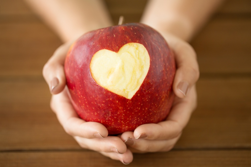Close up of hands holding ripe red apple with carved heart shape over wooden table