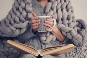 woman covered with a blanket reading a book and with a warm drink.